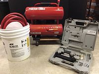 View of PorterCable air compressor, red air hose coiled next to a white bucket, and brad nailer carrying case.