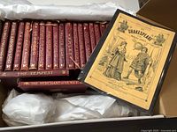 Box containing 22 small maroon leather bound volumes of Shakespeare plays with gold embossed titles, a similarly bound volume in cloth, a red leather bound volume of The Merchant of Venice, and a yellow booklet titled 'Shakespeare Boiled Down'