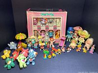 Full overview of the vintage Care Bears, Cabbage Patch Kids minis, Strawberry Shortcake figures, Monchhichis and accessories on dark fabric background.