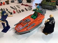 Photo showing orange armed boat vehicle with seated green-uniformed figure and two standing figures, along with weapons and accessories in background.