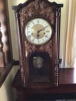 Full view of vintage wooden wall clock showing ornate carved floral details, clock face with Roman numerals, and pendulum with starburst design visible through glass panel.