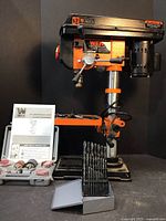 Overview of drill press on base with manual, sanding adapter kit and drill bit rack laid in front