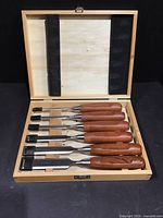 Six Wood River chisels with wooden handles in a fitted wooden box, showing the tools neatly held in place with some signs of wear on blades.