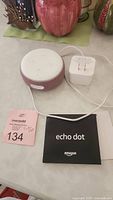 Photo of Amazon Echo Dot smart speaker in pinkish fabric color with white top, power adapter, and black Echo Dot instruction booklet on a table.