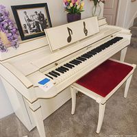 Front angle view of Baldwin Acrosonic spinet piano with custom white finish and matching red-padded bench showing keyboard, music stand with music note design, and legs.