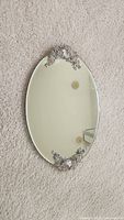 Oval vanity mirror lying flat on carpet showing rhinestone flower decoration on one end.