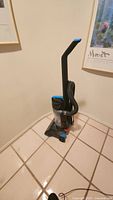 Full view of the upright vacuum cleaner standing upright on tiled floor, showing the handle, base, and hose assembly.