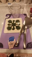 Photo showing the Allergen purple cutting board with handle, ceramic hotplate with leaf design, clear measuring cup with spout, and large metal masher placed on the cutting board.