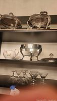 Shelf display showing 3 silverplate servers, ceramic teapot with lid, and 6 crystal dessert glasses beneath.