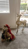 Santa figurine dressed in red velvet with fur sitting next to ornate metal reindeer on stand