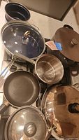Multiple pots and pans arranged on stovetop and countertop including navy blue ceramic, stainless steel pots, strainer, non-stick frying pan and lids