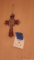 Photo of a red and silver decorative cross hanging on a chain against a wooden background with a MaxSold tag.