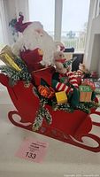 Front angle of red wooden Santa sleigh with Santa and elf dolls, wrapped gifts, Christmas greenery and wooden blocks.