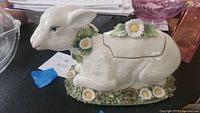 Side view of the vintage ceramic lamb box showing the lamb with daisies on its neck and base, highlighting the slot of the hidden compartment on the back.