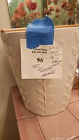Close-up photo of a white ceramic bucket wastebasket showing embossed leaf stem patterns and scalloped top edge with natural rim color. A label is attached with some text and blue tape.