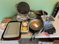 Overview of assorted bakeware and cooking pans on table including fry pans, mesh basket, ceramic pie plate, baking pans, pizza tray, and waffle iron