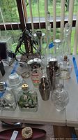 Photo showing multiple glass decanters, two metal cocktail shakers, dark metal wire wine rack, and assorted glass bottles.