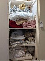 Multiple folded and rolled bed linens on shelves including various blankets, comforters, sheets, pillowcases in neutral and pastel colors.