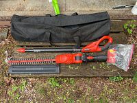 Complete trimmer parts laid out on wooden bench including red and black motor and handle, extension pole, blade, battery, charger, and carrying case.