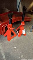Two red adjustable metal jack stands with black threaded supports standing upright on a concrete floor, showing use signs and some rust patches.