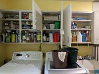 Wide view of cabinets above washer and dryer containing multiple bottles of cleaning products along with buckets on dryer.