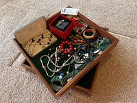 Wooden jewelry trays filled with assorted costume jewelry including necklaces, bracelets, brooches, earrings, and a red beaded bracelet.
