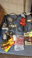 Overview of electric drills, heat gun, testers, and various accessories on a blue table surface, showing cords and packaging.