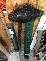 View of rolls of fencing including green plastic coated metal fencing, black deer fencing netting, and a roll in black wrapping inside a shed.