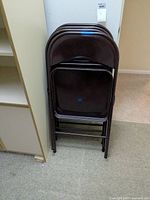 Four stacked Meco folding chairs in dark brown, standing upright against wall.