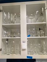Full view of cabinet with multiple shelves filled with varied glassware including tall glasses, stemmed glasses, cordials, shot glasses, and other barware.