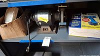 Photo of Alltrade 1/2 HP bench grinder with power cord and extra brushes on shelf alongside original box packaging.