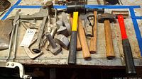 Overview of all hand tools on wooden surface including clamps, vise grips, several hammers, mallets and axe head.