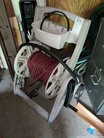 White plastic hose reel with maroon garden hose wrapped around and an extra black hose over top, stored indoors on a shed floor.