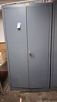 Front view of the tall gray metal cabinet with two doors closed and a tag attached.