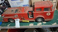 Front and side view of the red metal toy fire truck showing Texaco logo, rust, and missing parts on a green surface with boxes in the background.