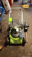 Full view of the Greenworks electric lawn mower with handle and hedge trimmer placed beside it on the floor