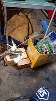 Wide photo showing folded tarps, yellow box with painting supplies, manual hand sprayer, paint rollers and brushes on concrete floor.