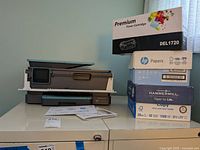 Full view of HP OfficeJet Pro 8028 printer on filing cabinet with paperwork and three reams of paper stacked next to a premium toner cartridge box labeled DEL1720.