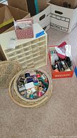 Overview of sewing basket filled with various colored thread spools and notions, with a multi-drawer organizer beside it on the floor.