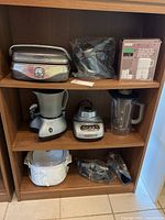 Shelf with all six appliances including Calphalon waffle maker, Cuisinart Smart Stick parts, Zeico coffee maker box, Back to Basics cocoa-latte maker, KitchenAid blender jar and base, and CrockPot slow cooker