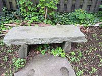 Front view of the stone bench set outdoors showing the bench seat and visible stone legs, surrounded by greenery and positioned on soil with a large flat stone slab in front.