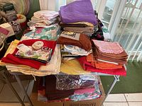 Table covered with folded assorted table linens including napkins, cloths, and towels in various colors, some tagged with price labels