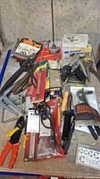 Wide arrangement of assorted tools including wire strippers, clamps, scales, and boxed hardware supplies on concrete surface.