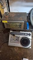 Photo showing both the rusty metal RMA radio on top and the GE cassette player below, with visible wear and industrial surroundings.