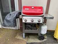 Front-left view of red Weber grill with cover draped, showing three control knobs and side tables