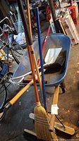 Overall view of the metal wheelbarrow holding several brooms and shovels in a garage setting.