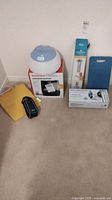 Overall view of all items included in the lot showing 8 distinct products in their boxes or packaging including humidifiers, blood pressure monitor, bed alarm system, kneeling pad and bath bar.
