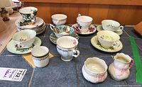 Collection of teacups and saucers in varied floral and decorative patterns in pastel colors, along with a small cylindrical cup, all arranged on a table.