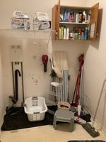 View of the cleaning supplies including brooms, laundry basket, mop, step stool, washboard, and wooden cabinet with various cleaning liquids and sprays.