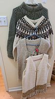 Group of four sweaters hanging on wall including a green turtleneck, cream V-neck with pattern, beige sweater, and sleeveless vest.